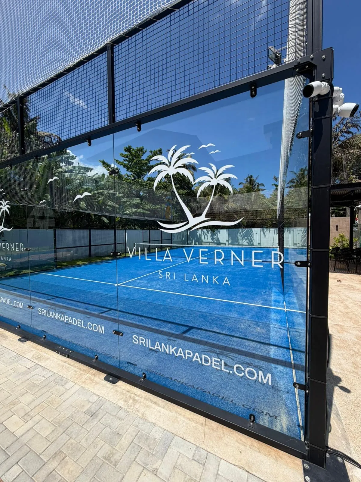 The padel court's glass wall with the Villa Verner logo, the blue court behind