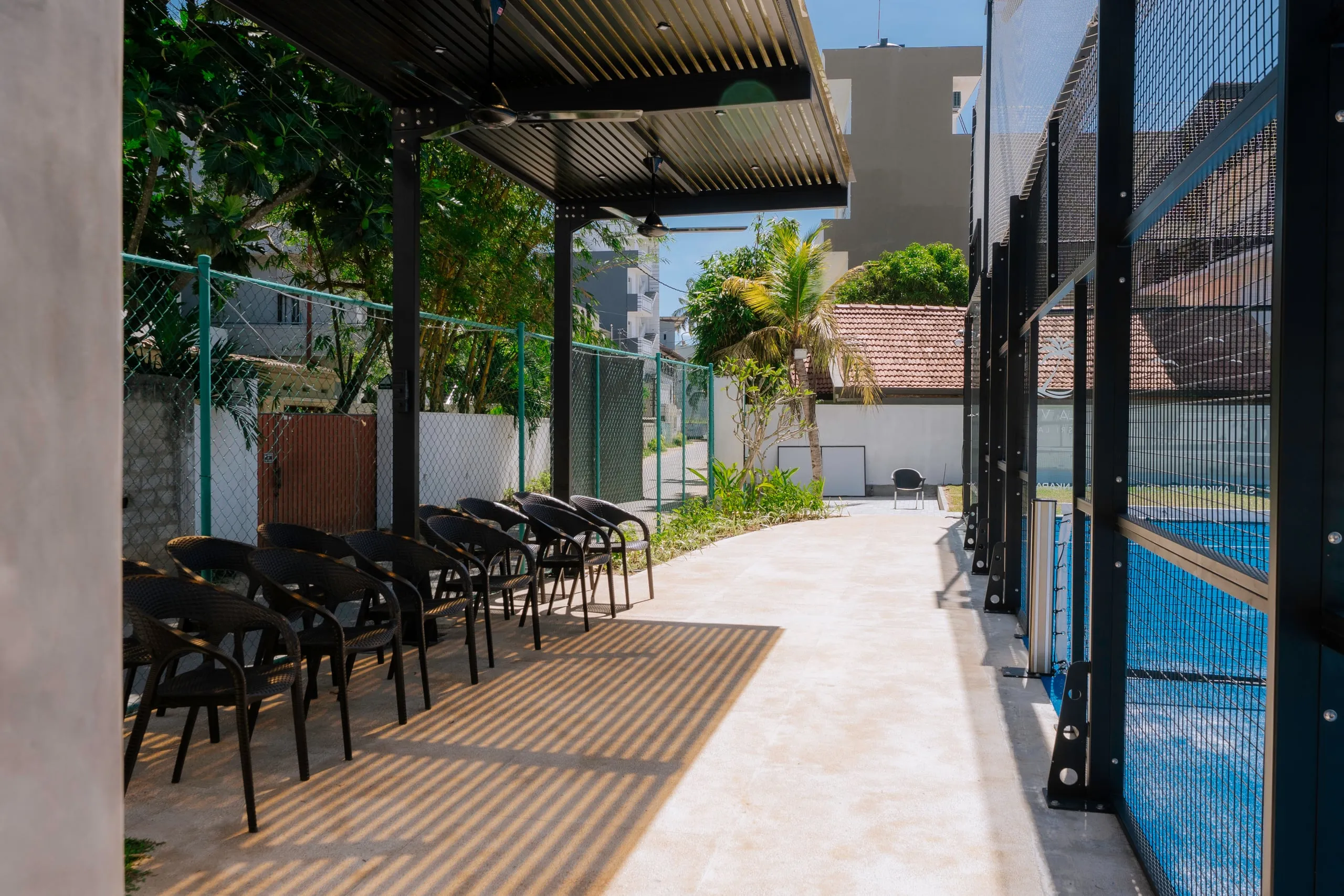A pergola with black chairs beside the padel court, palms and neighbouring houses behind
