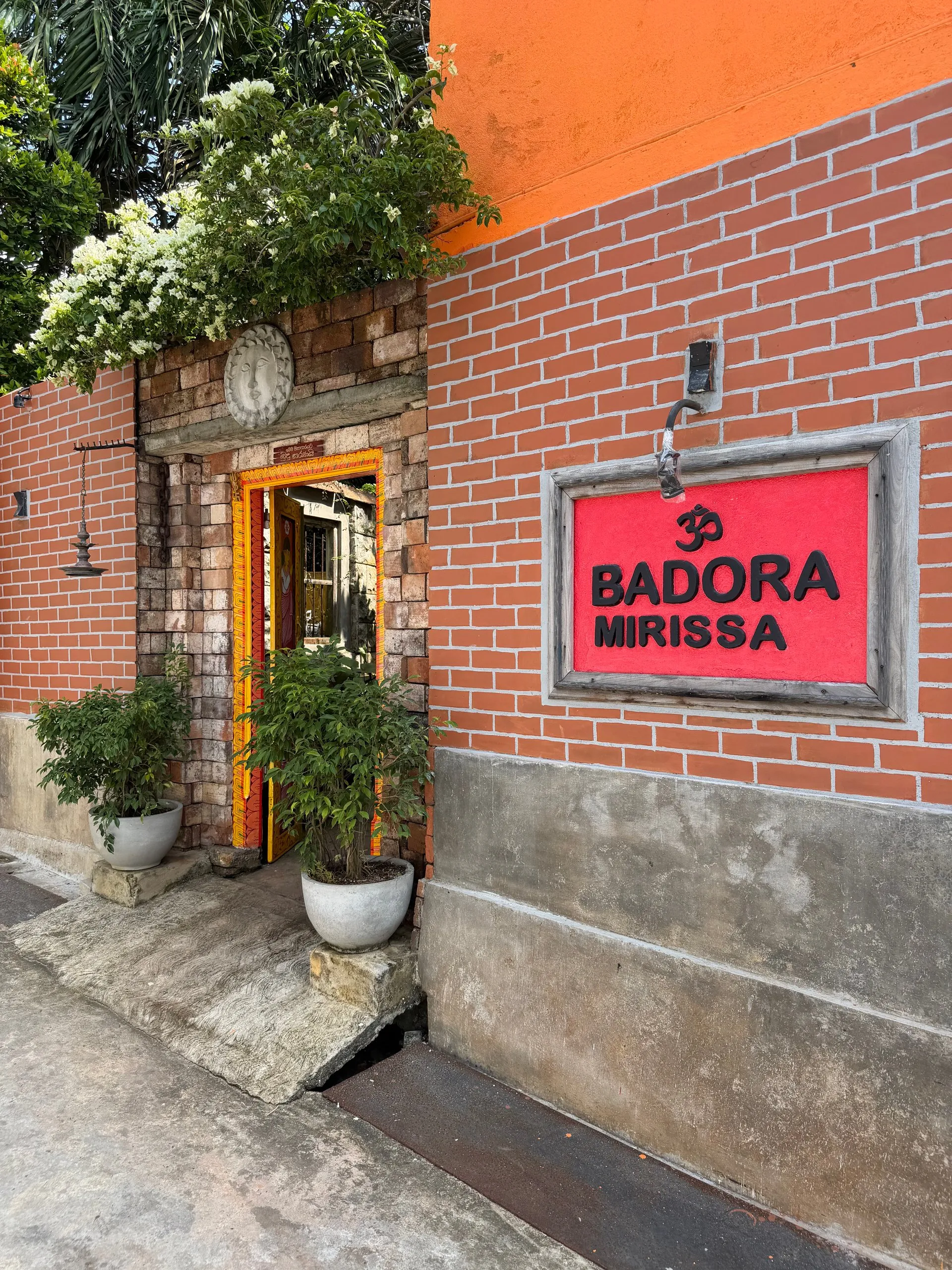 Badora Spa sissepääs Mirissas — punane silt telliskiviseinal ja saialilledega kaunistatud kaaruks