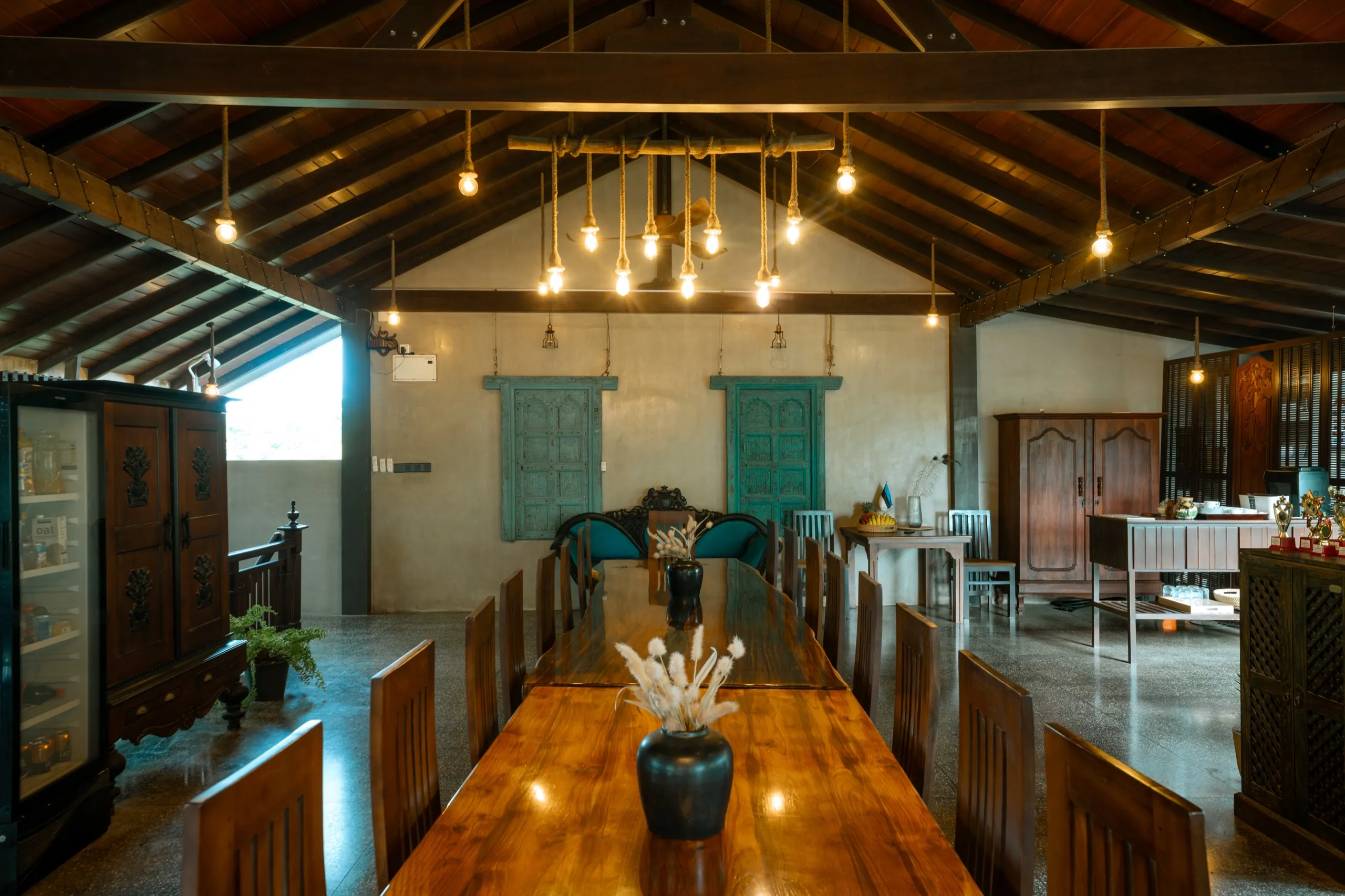 A long timber dining table with chairs under filament bulbs, turquoise window shutters behind
